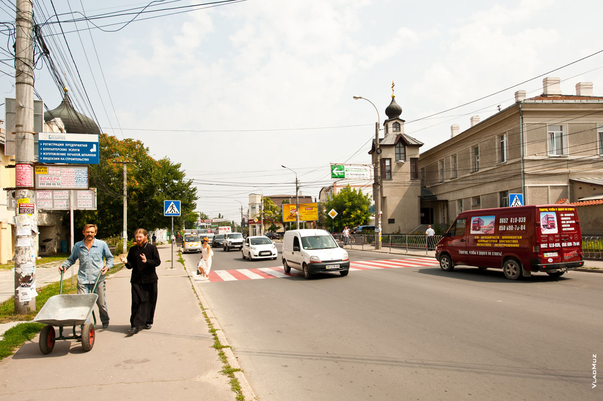 Фото мужчины с тачкой и священника в черном, идущих по улице Кавказской в Симферополе, справа видна часовня святителя Луки