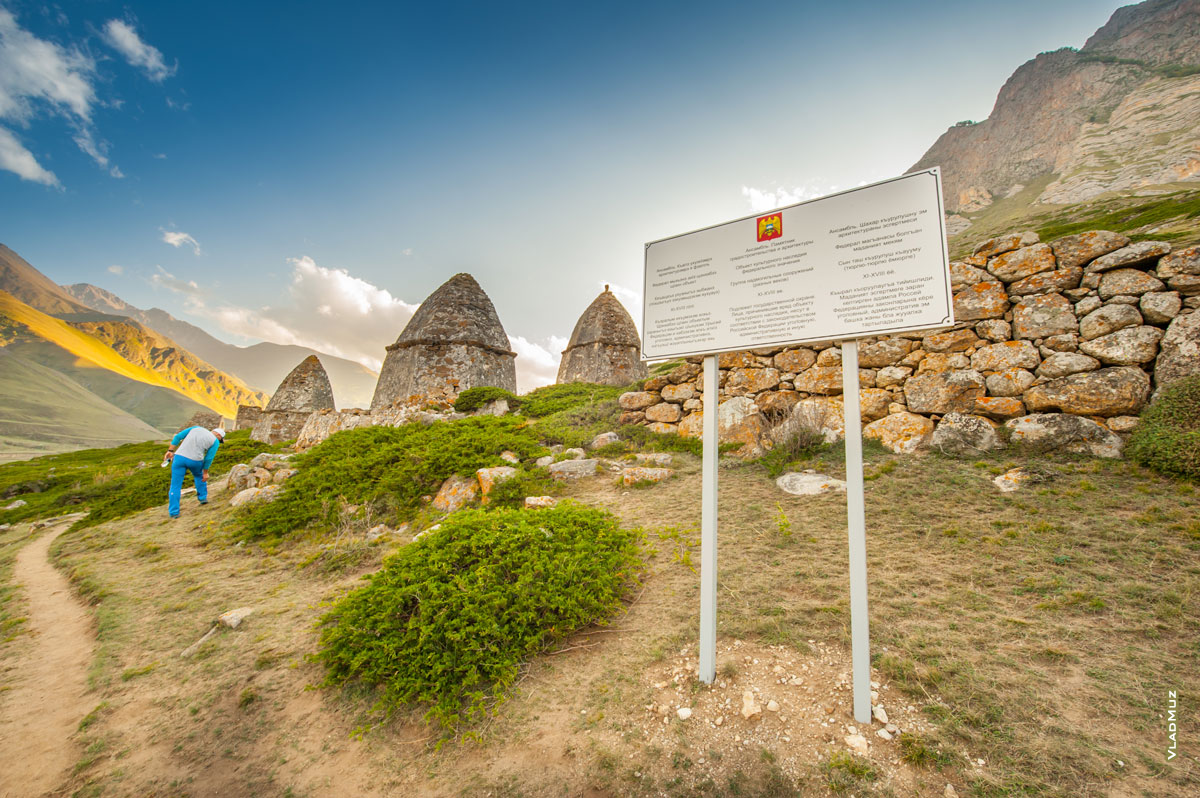 Фото щита с информацией о могильниках Фардык-Кешене на фоне «Города мертвых» в Эльтюбю