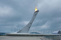 Фото горящего Олимпийского огня «Сочи 2014» на безлюдной Олимпийской площади в парке. Вдали — Большой ледовый дворец