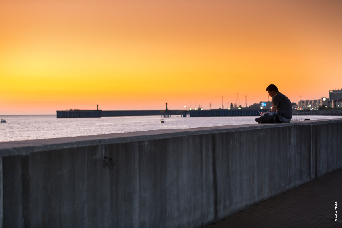 Фото мужчины с телефоном на парапете набережной на фоне Имеретинского морского порта, после заката солнца