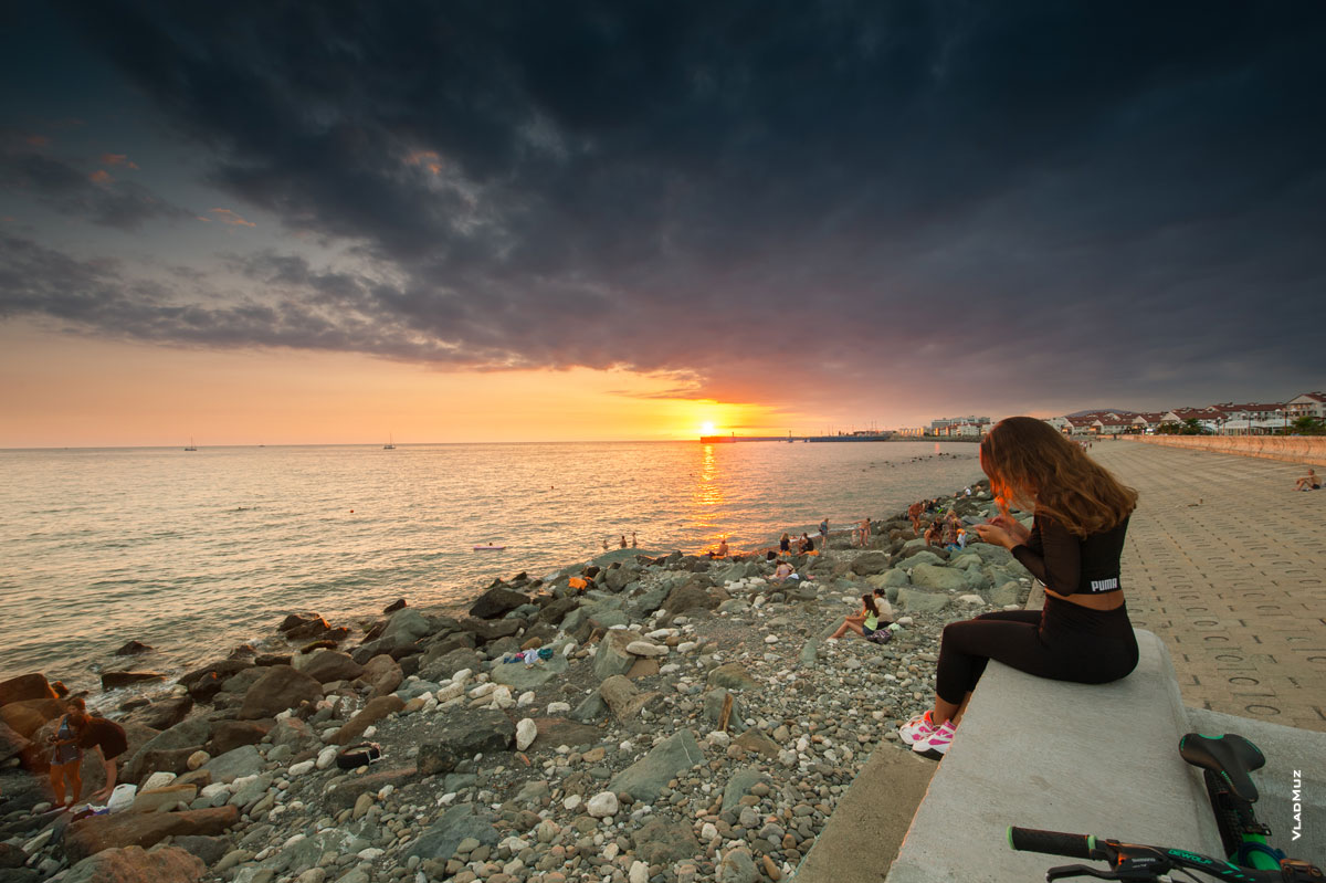 Фото девушки с телефоном с видом на море, закат солнца и Имеретинский городской пляж. Летний морской фотопейзаж